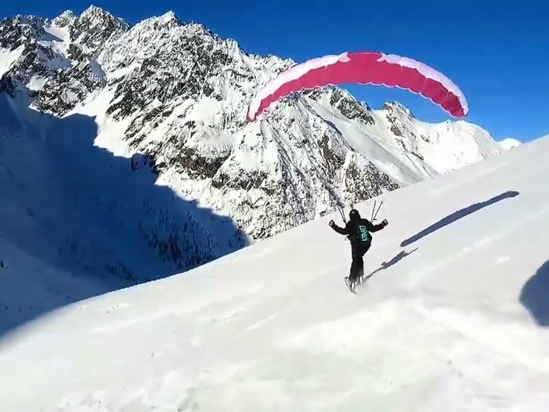 Parapentiste avec voile rose déployée en train de décoller ou atterrir sur une pente enneigée en montagne.