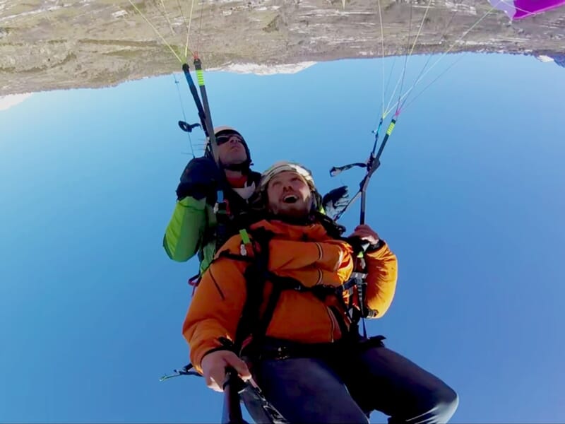 Deux personnes en tandem font un baptême de parapente au-dessus d'un paysage montagneux sous un ciel bleu clair.