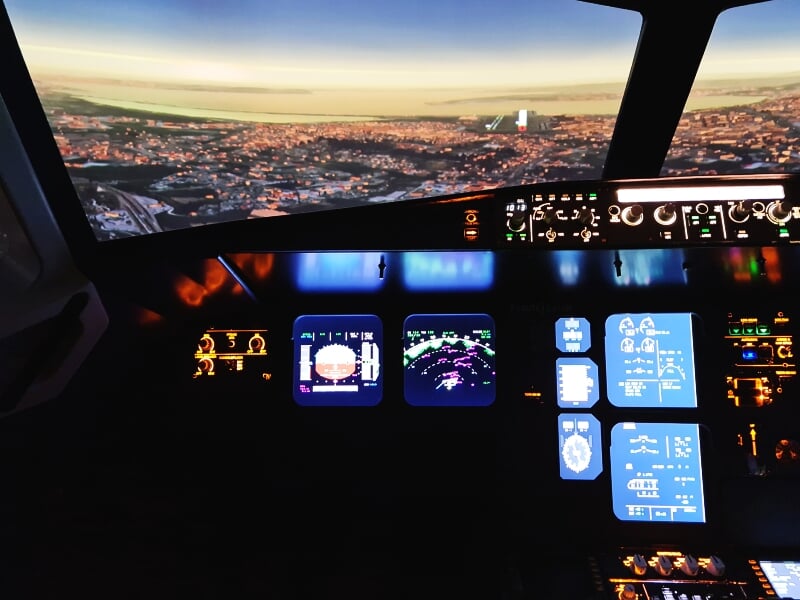 Tableau de bord lumineux d'un simulateur de vol avec écrans et commandes, vue sur la ville au loin.