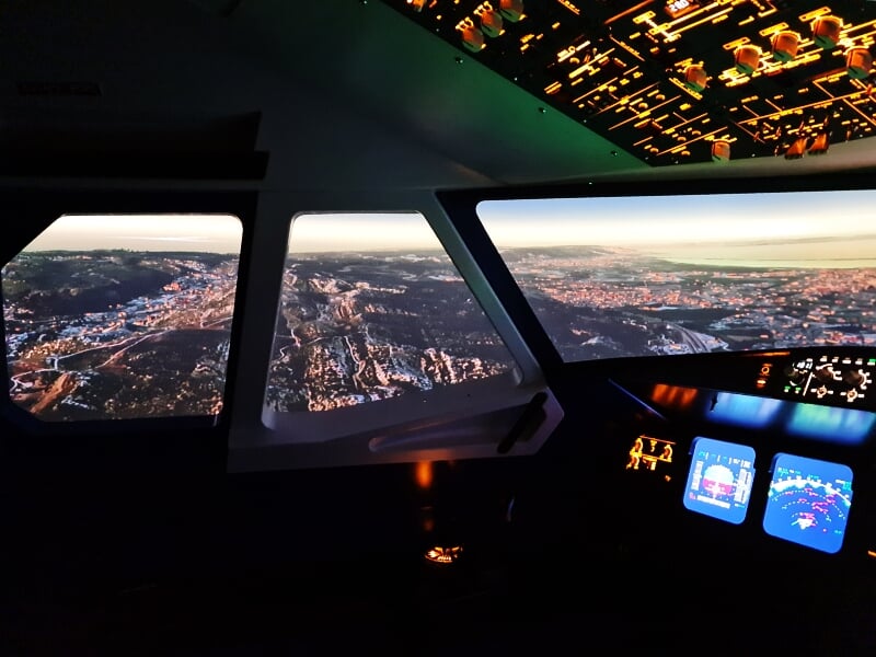 Vue depuis le cockpit d'un simulateur de pilotage avec instruments et paysage urbain au coucher du soleil.