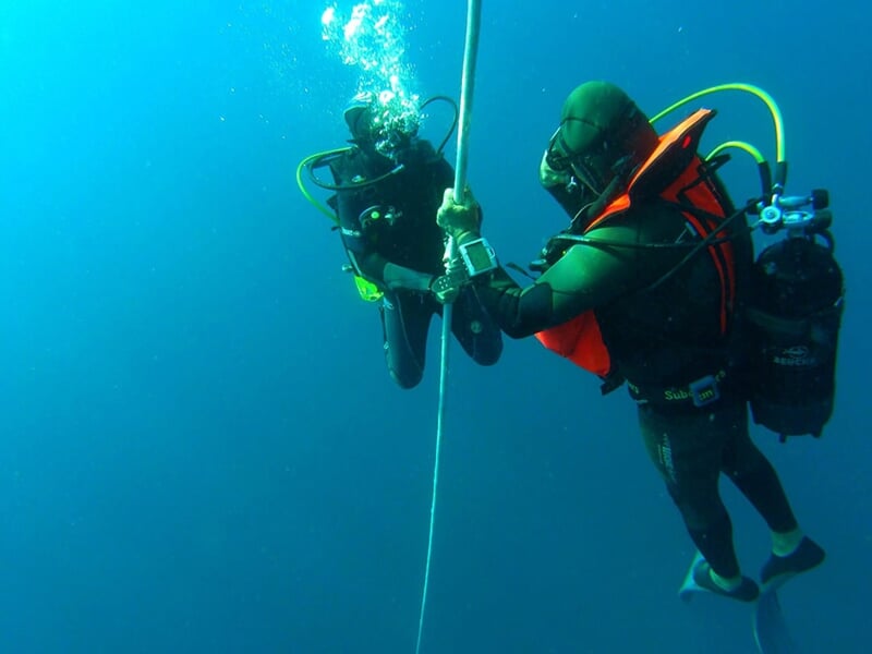 Deux plongeurs en combinaison noire et gilet orange remontant le long d'une corde sous l'eau bleue profonde.
