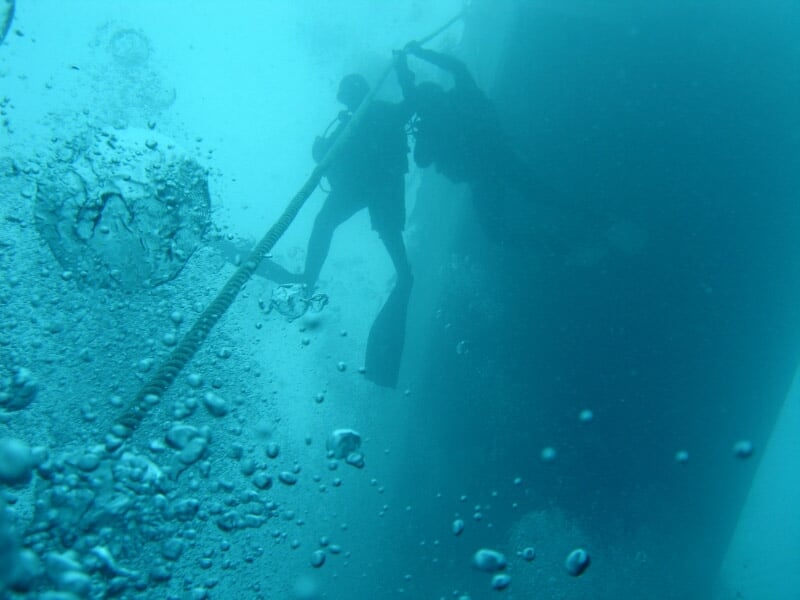 Plongeurs sous l'eau tenant une corde, entourés de bulles, avec un grand objet sombre en arrière-plan.
