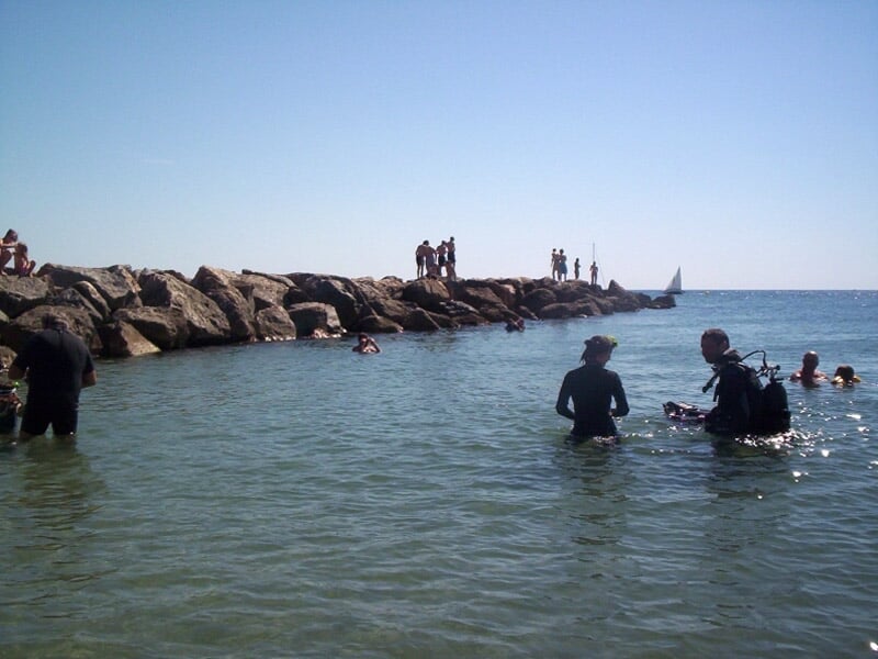 Des plongeurs et baigneurs dans une eau peu profonde près d'une jetée rocheuse sous un ciel clair.