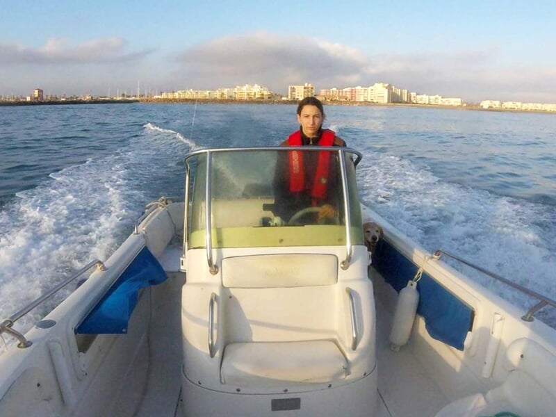 Personne au volant d'un petit bateau blanc, portant un gilet de sauvetage rouge, en mer calme.
