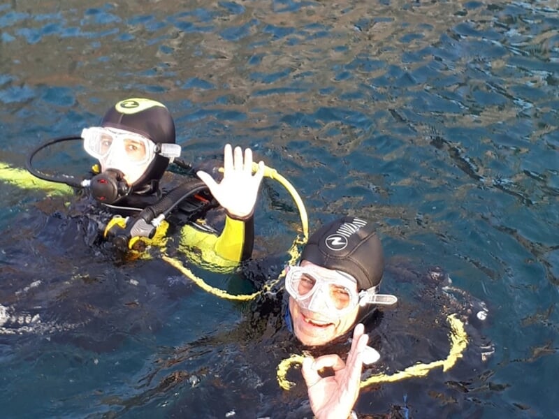 Deux plongeurs souriants en surface de l'eau, équipés de combinaisons et masques de plongée, saluent de la main.