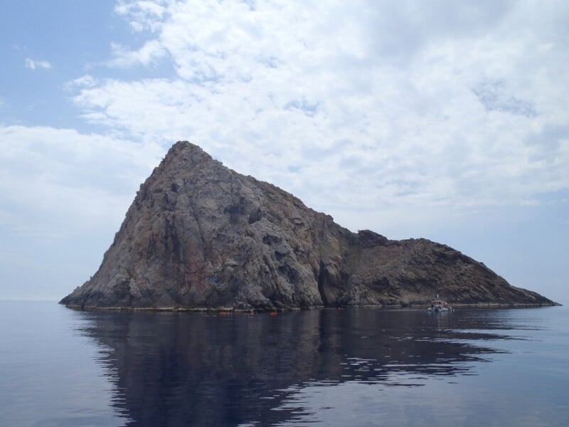 Île rocheuse isolée se reflétant dans une mer calme sous un ciel nuageux, sans présence humaine visible.