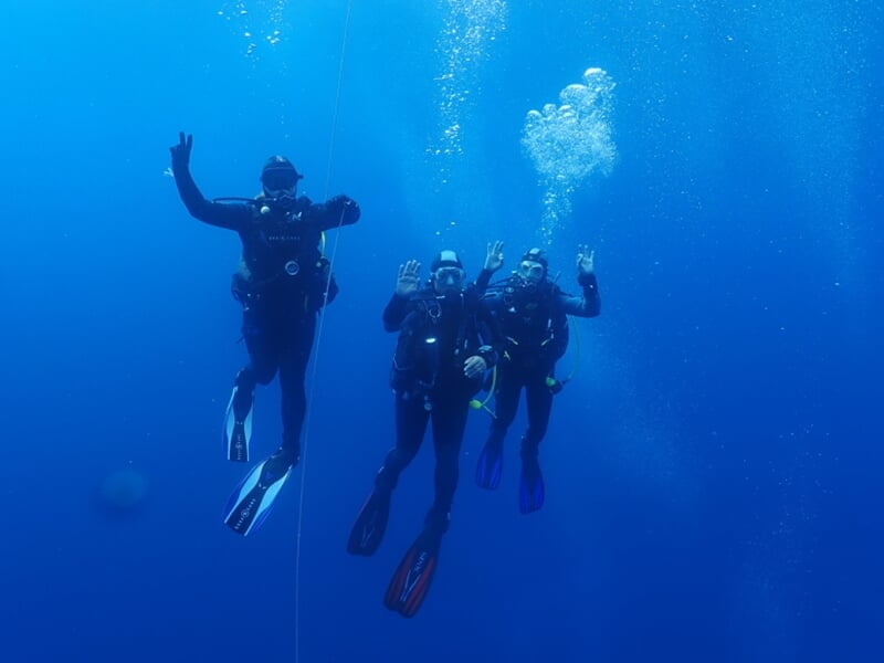 Quatre plongeurs sous l'eau, en combinaison noire, font des signes de la main et flottent dans une eau bleue.