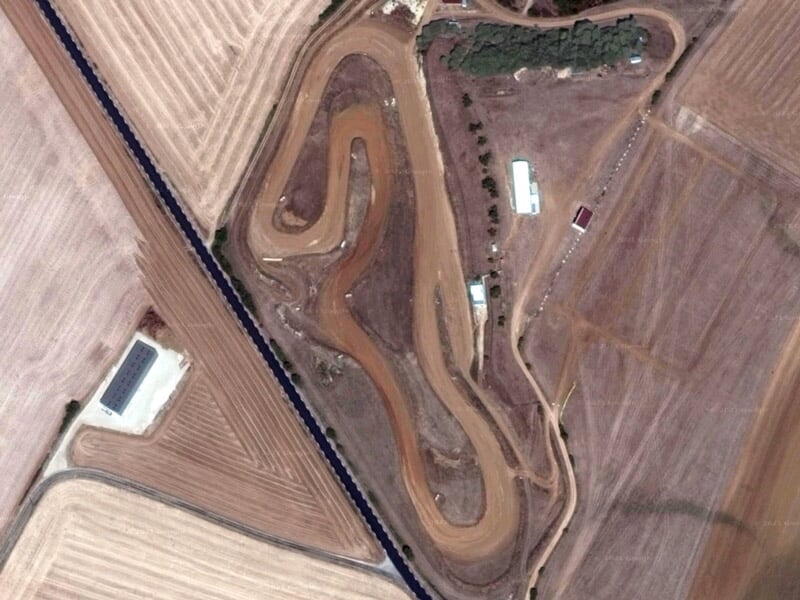 Vue aérienne d'un circuit de rallye terre sinueux entouré de champs agricoles et de bâtiments.