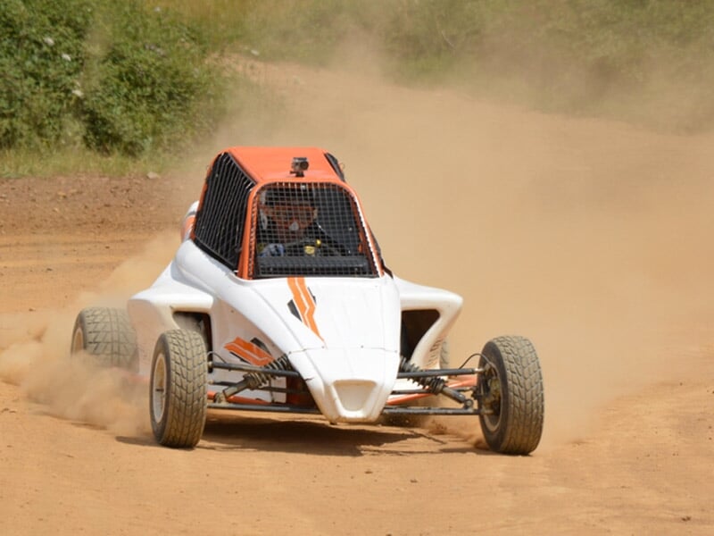 Buggy blanc et orange sur piste terre, en virage avec poussière soulevée derrière le véhicule.