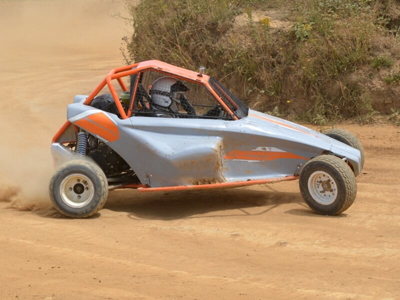 Buggy gris et orange sur circuit terre, en virage avec poussière soulevée sur piste de rallye.