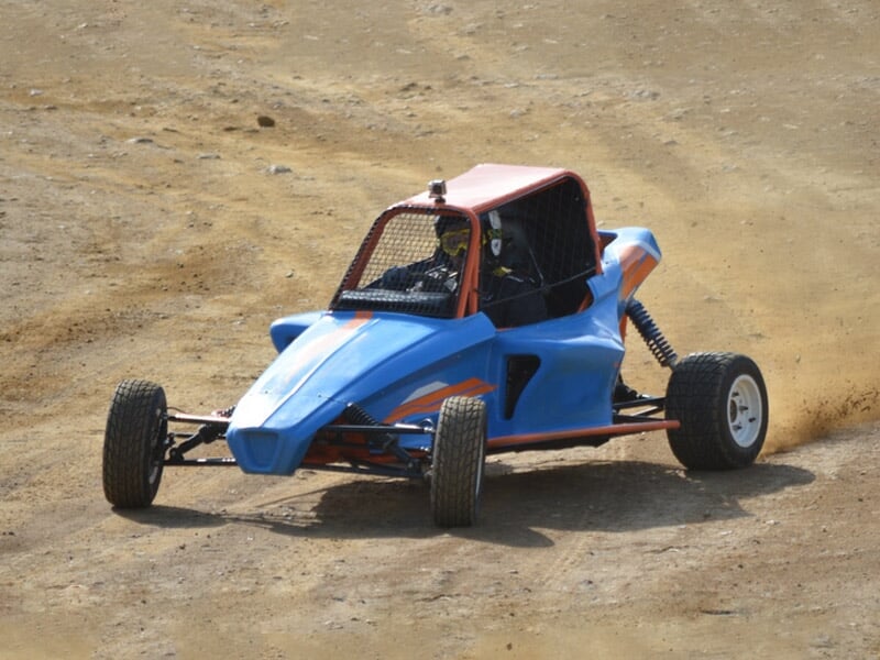 Buggy bleu sur piste terre, en virage avec conducteur casqué, poussière soulevée autour.