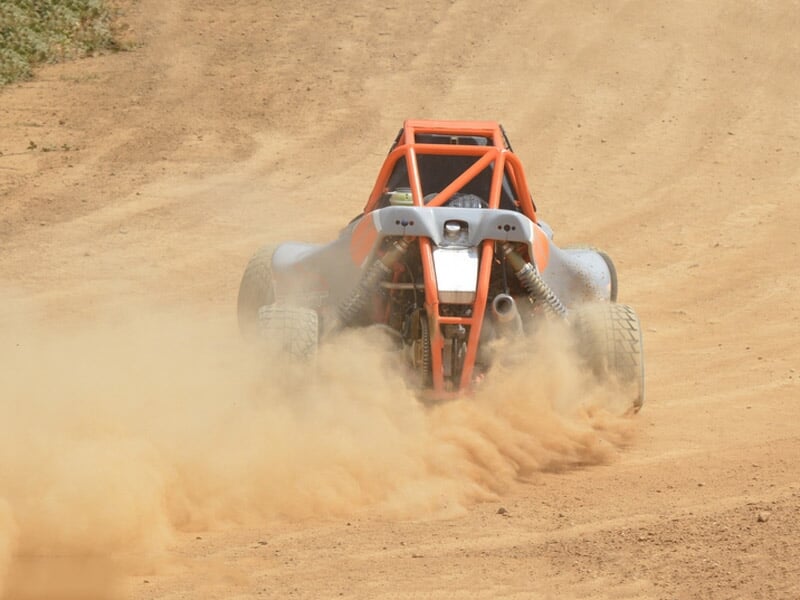 Buggy orange sur circuit terre, vu de face, soulevant un nuage de poussière en pleine course.