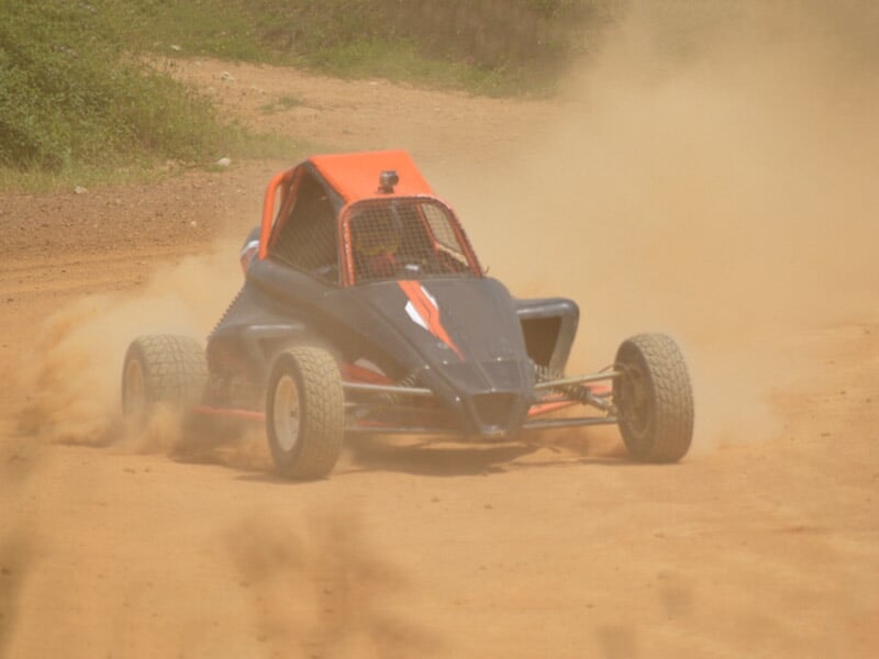 Buggy noir et orange sur circuit terre, en virage avec poussière soulevée, ambiance rallye.