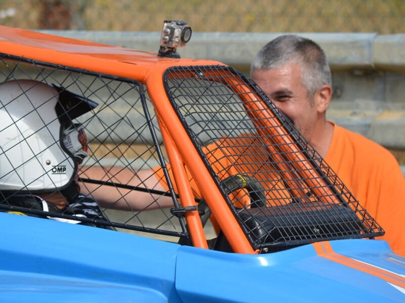 Pilote casqué dans buggy bleu, discutant avec un homme en t-shirt orange près du circuit.