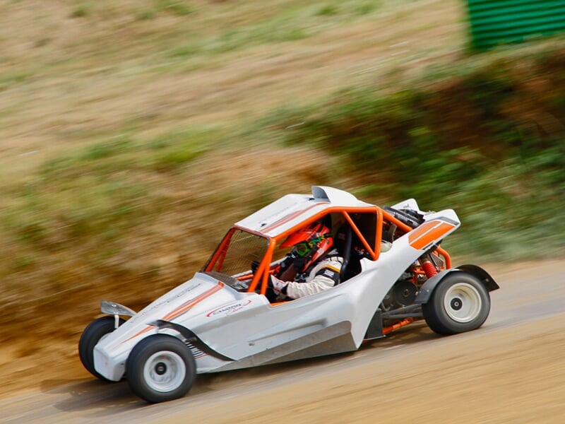 Buggy blanc et orange sur circuit terre, en virage rapide, poussière soulevée sur piste.