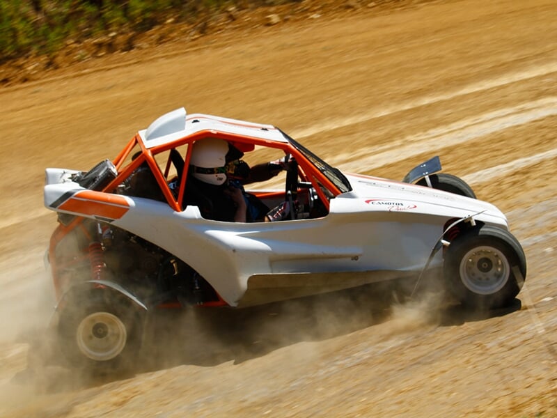 Buggy blanc et orange sur piste terre, en virage serré, pilote casqué concentré au volant.