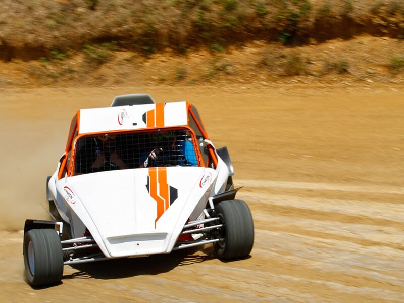 Buggy blanc et orange sur circuit terre, en virage avec poussière soulevée, ambiance sportive.