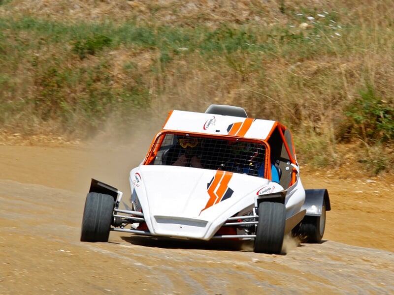 Buggy blanc et orange sur piste terre, en virage serré, pilote casqué visible au volant.