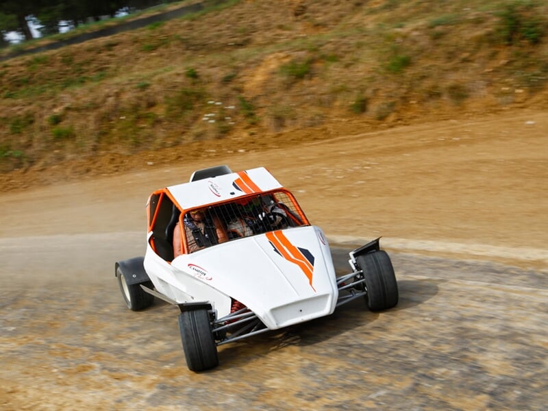 Buggy blanc et orange sur circuit terre, en virage rapide, poussière soulevée sur la piste.