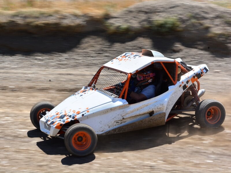 Buggy blanc et orange sur piste terre, en virage serré, pilote casqué concentré au volant.