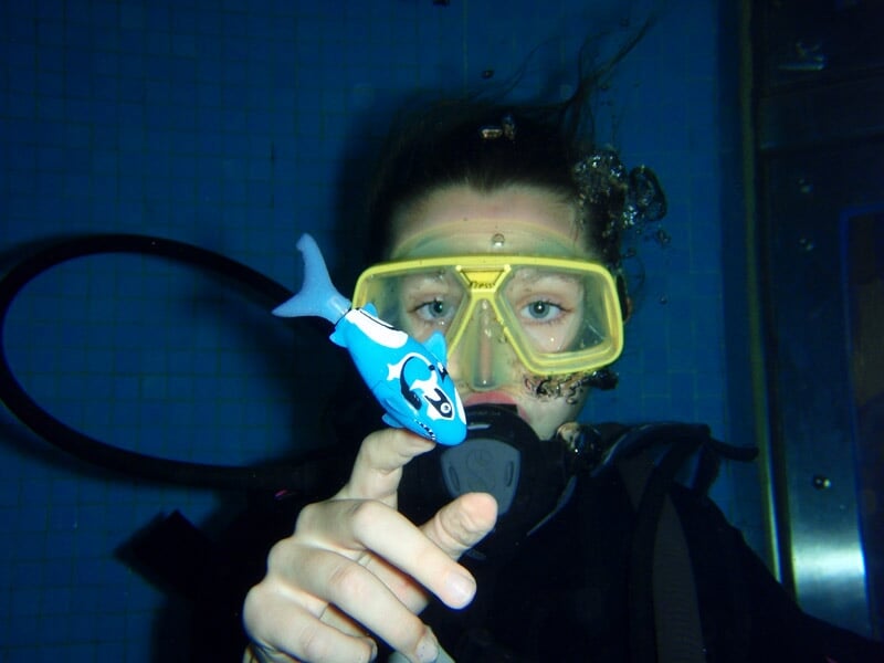 Plongeur sous l'eau portant un masque jaune et tenant un jouet poisson bleu, avec bulles autour.