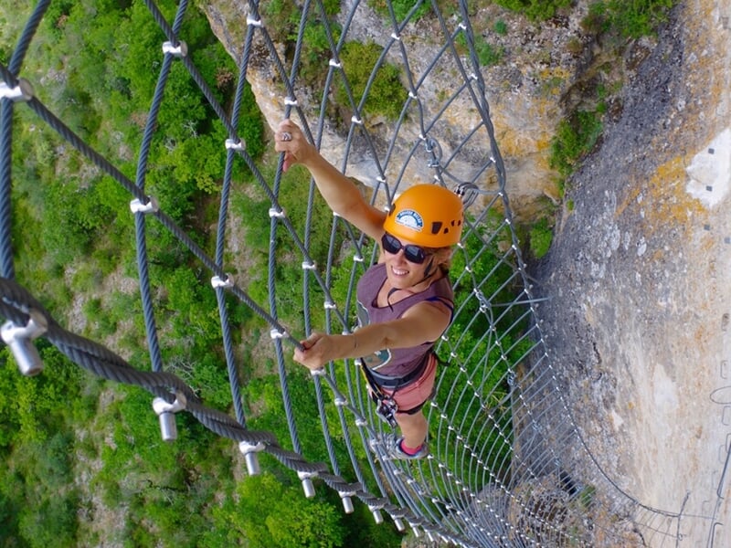 Escaladeuse souriante portant un casque jaune et des lunettes de soleil, grimpant sur un filet de corde suspendu au-dessus d'un précipice verdoyant.