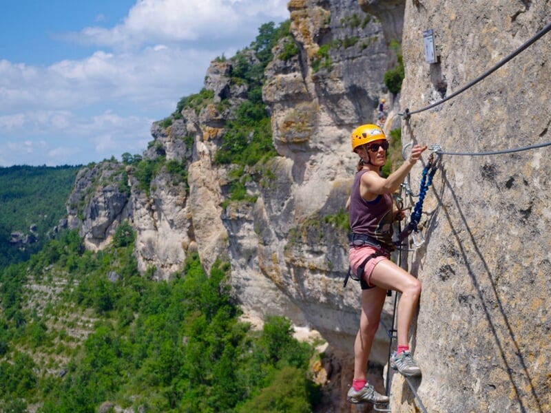 Escaladeuse avec casque jaune et harnais, progressant sur une via ferrata contre une falaise rocheuse, avec une vue sur la vallée en arrière-plan.