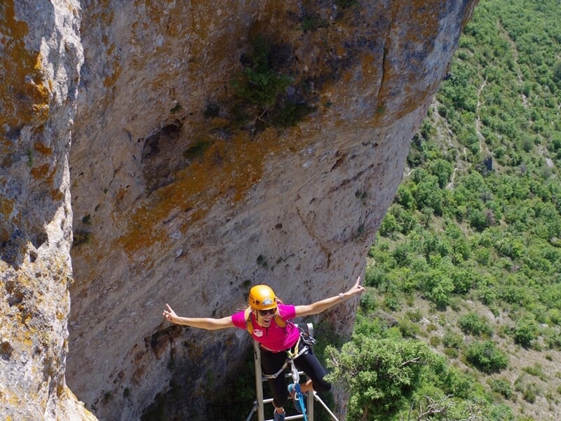 Personne en tenue de sécurité jaune et rose pratiquant la via ferrata sur une paroi rocheuse en France.