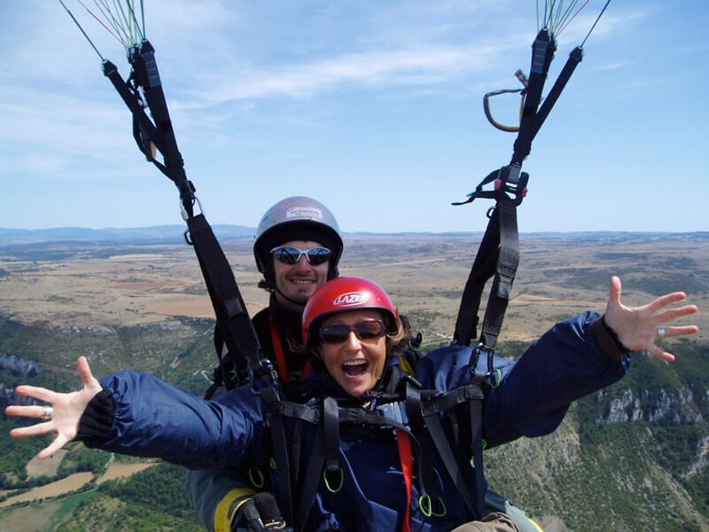 Duo en parapente en plein vol, avec la passagère souriante et les bras ouverts, survolant un paysage vallonné.