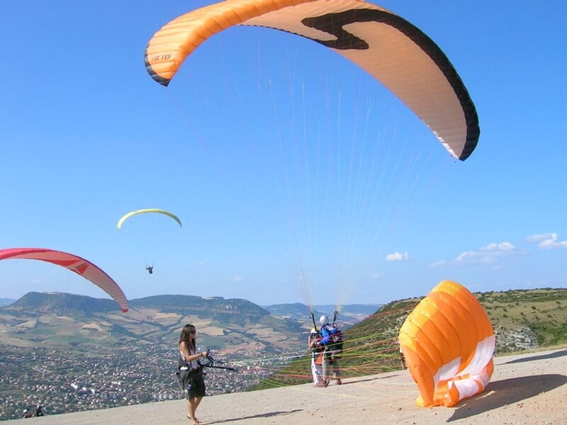 Parapentiste sous une voile orange prêt à décoller d'une colline, avec plusieurs autres parapentes en vol au-dessus d'une vallée.