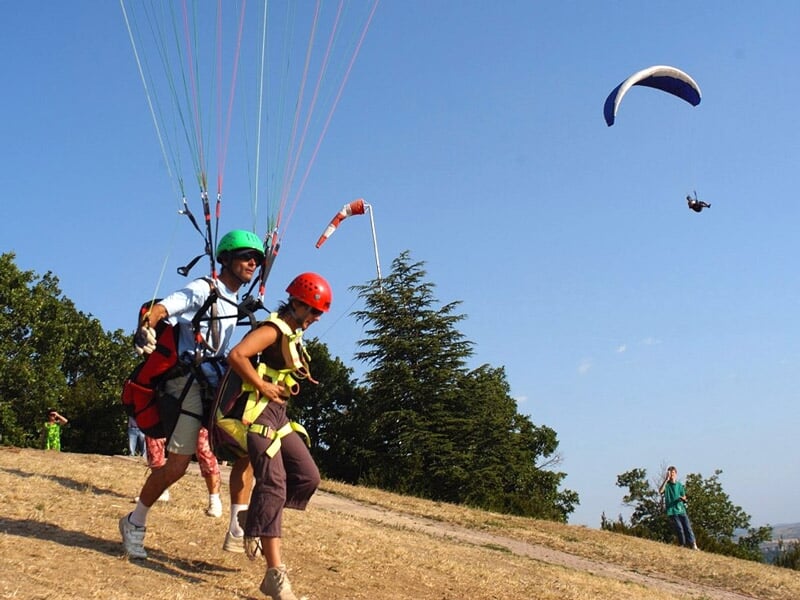 Duo en parapente courant pour décoller d'une colline sous un ciel bleu, avec un autre parapente en vol en arrière-plan.