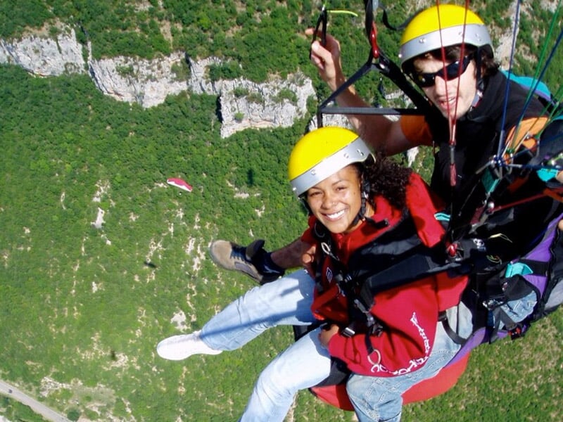 Duo en parapente survolant une vallée verdoyante, avec la passagère souriante portant un casque jaune.