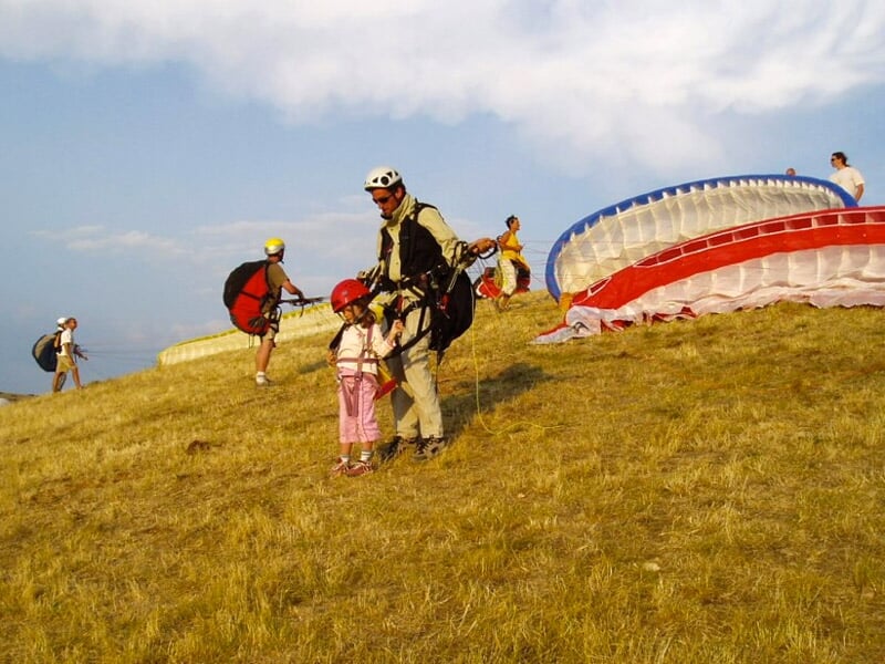 Enfant portant un casque et un harnais, accompagné d'un instructeur, préparant un décollage en parapente sur une colline herbeuse.