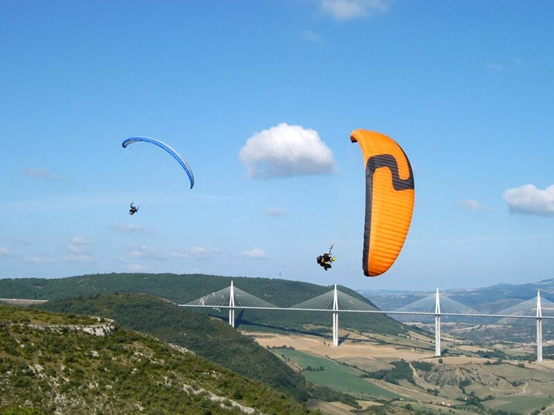 Deux parapentes, un bleu et un orange, volant au-dessus du viaduc de Millau et des paysages vallonnés.
