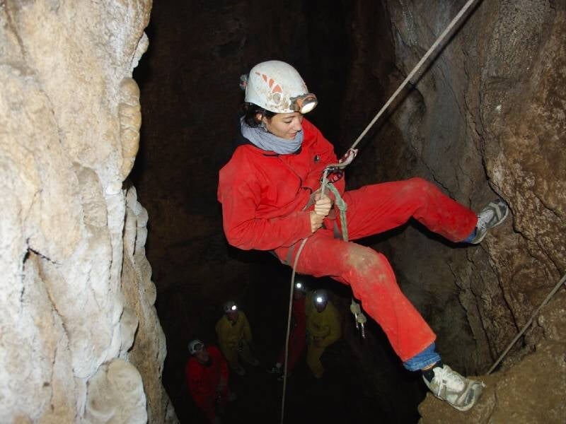 Spéléologue en combinaison rouge descendant dans une grotte sombre avec un casque et un harnais.