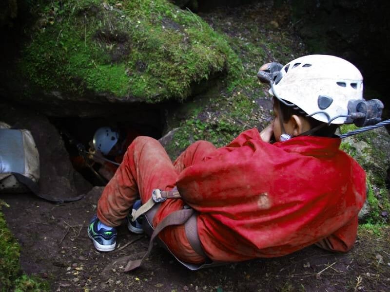 Spéléologue en combinaison rouge et casque blanc s'apprêtant à entrer dans une grotte, entouré de végétation.
