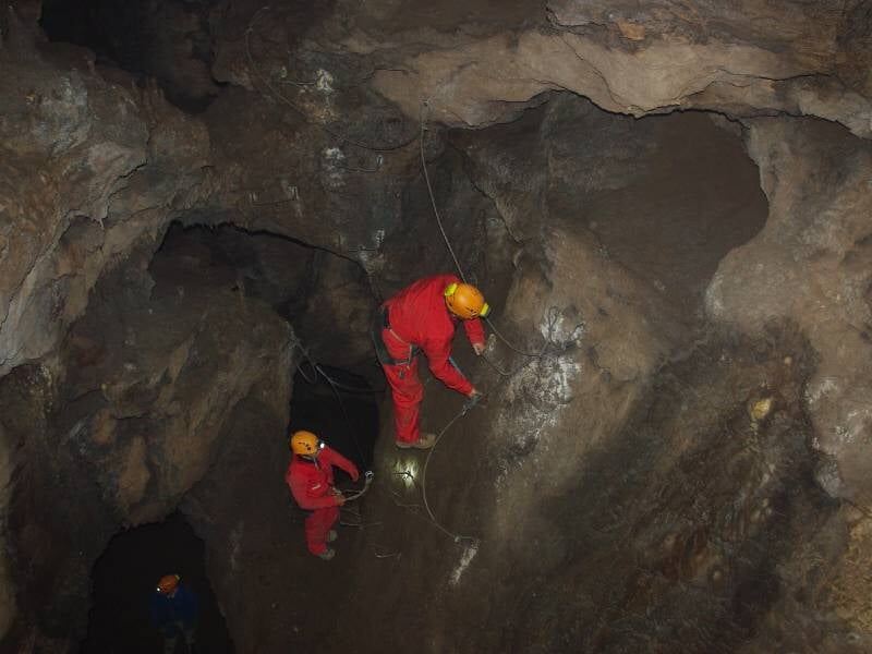 Spéléologues en tenue rouge et casque escaladant une paroi intérieure de la grotte, utilisant des cordes de sécurité pour progresser dans un passage étroit et sombre.