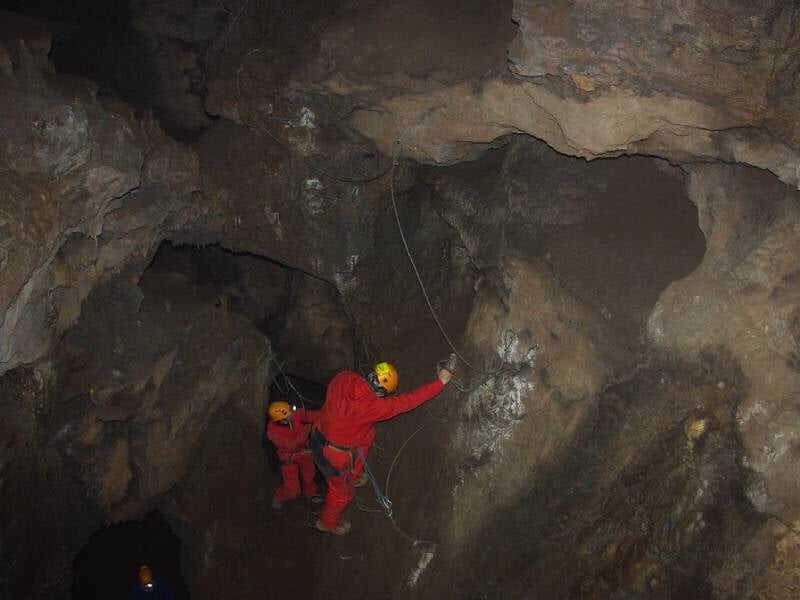 Spéléologues escaladant une paroi rocheuse à l'intérieur d'une grotte, attachés à des cordes de sécurité, portant des casques et combinaisons rouges.