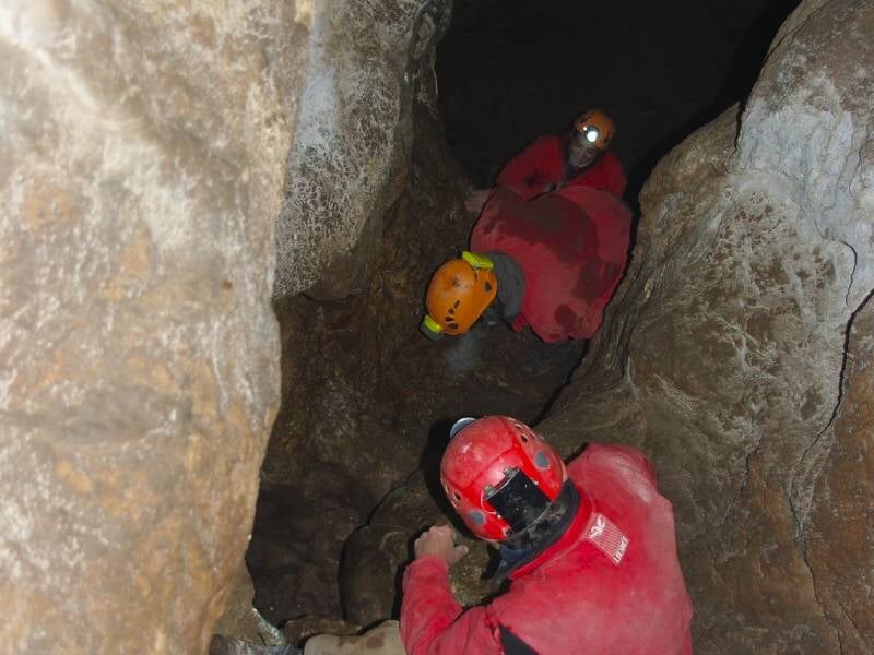 Groupe de spéléologues progressant dans un passage étroit et rocheux, portant des casques avec lampes frontales.