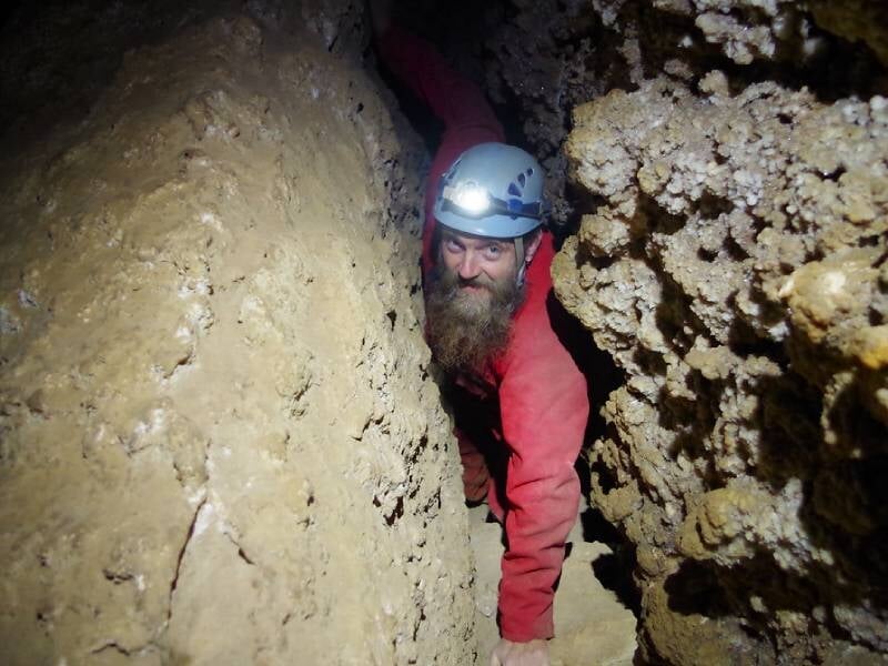Homme barbu en casque et tenue de spéléologue, rampe à travers un passage étroit dans une grotte, éclairé par une lampe frontale.