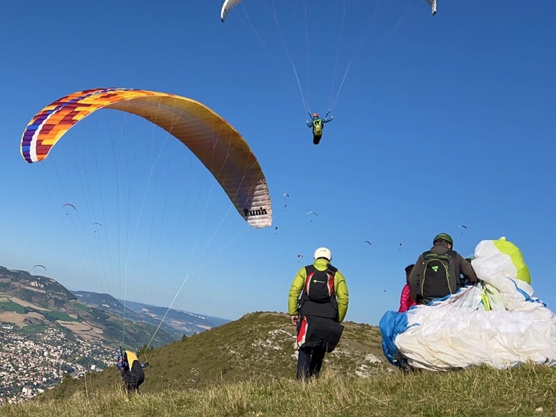 Deux personnes préparant leurs parapentes sur une colline, avec un parapente multicolore s'élevant dans le ciel bleu.