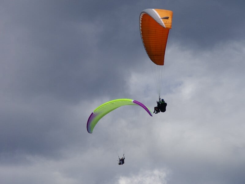 Deux parapentes en plein ciel nuageux, un vert et violet et un orange, avec des parapentistes suspendus.