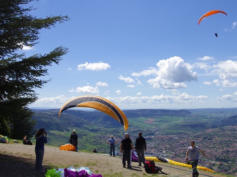 Plusieurs personnes se préparant pour le parapente sur une colline surplombant une vallée verdoyante, avec un parapente orange en plein vol.