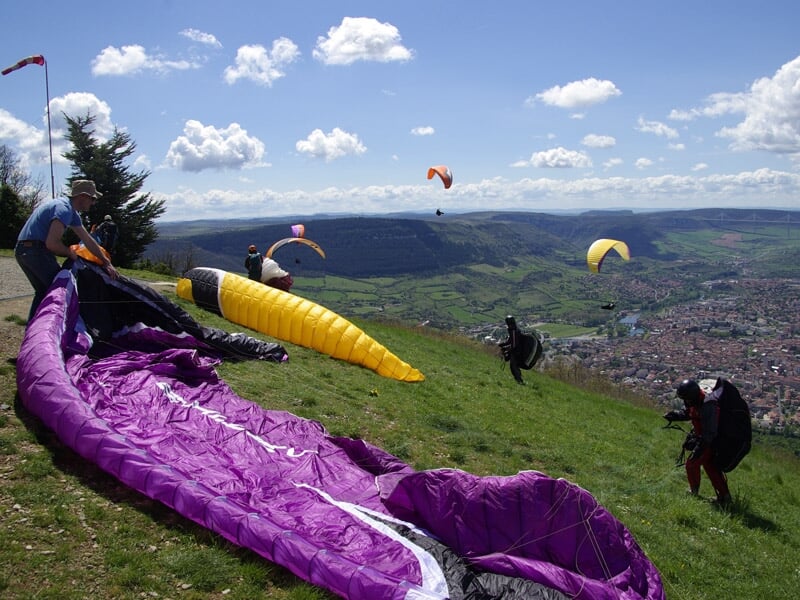 Préparation des parapentes colorés sur une colline verte avant le décollage, avec plusieurs parapentes déjà en vol en arrière-plan.
