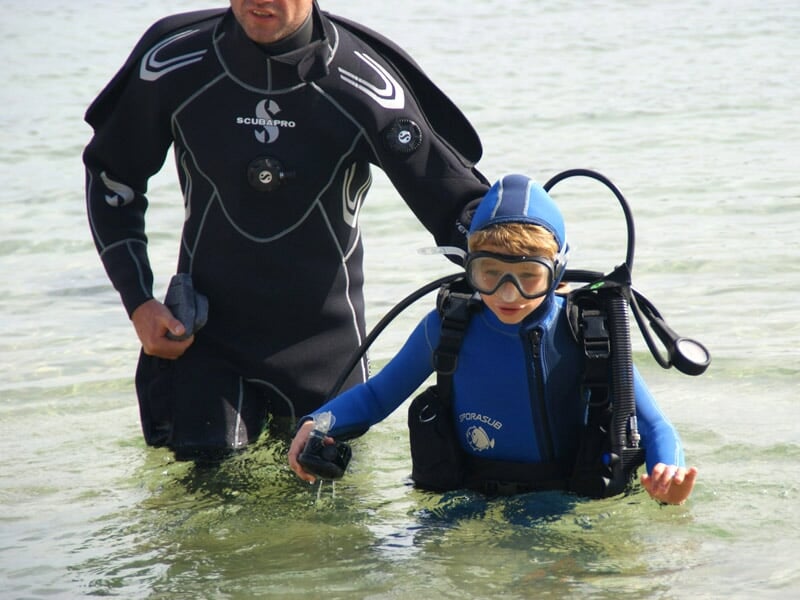 Un enfant en combinaison bleue et masque de plongée accompagné d'un adulte dans l'eau peu profonde.