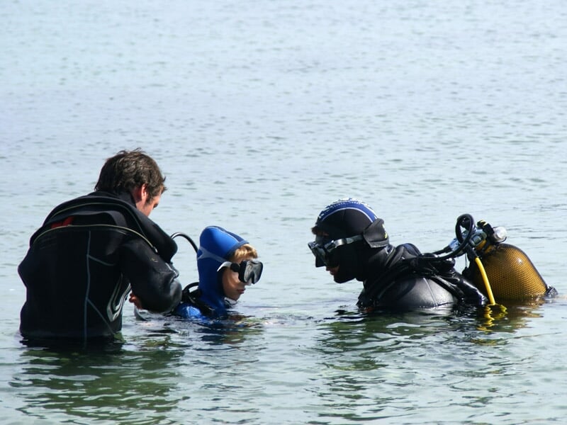 Deux adultes en combinaison noire encadrant un enfant en combinaison bleue dans l'eau pour une initiation à la plongée.