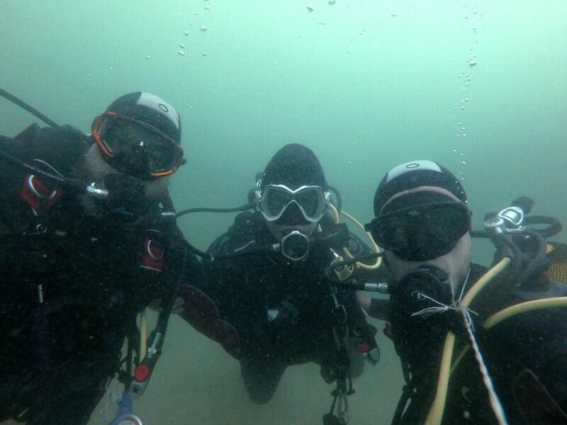 Trois plongeurs en combinaison noire posent pour une photo sous l'eau dans une eau légèrement trouble.