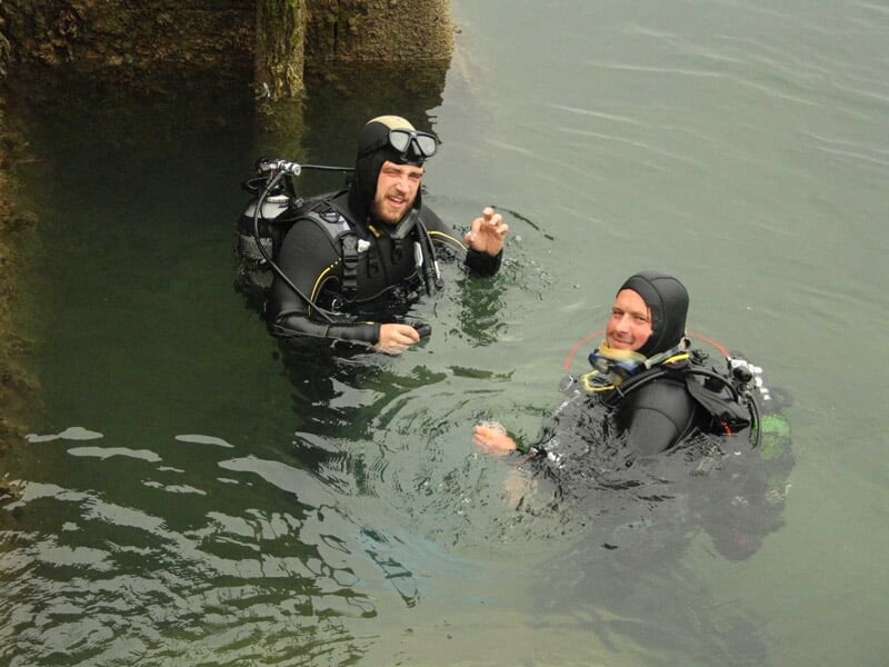 Deux plongeurs en combinaison noire s'entraînent dans l'eau près d'un mur en pierre, lors d'un baptême de plongée.