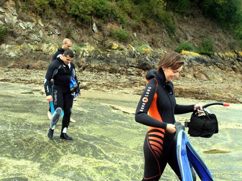 Trois personnes en combinaison de plongée marchant sur un rocher près de la mer, prêtes pour un baptême de plongée.