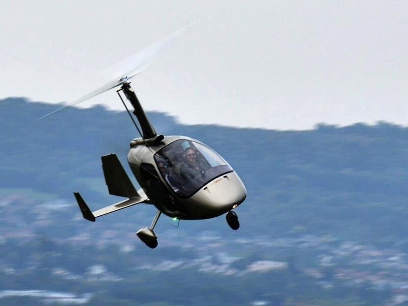Gyrocoptère noir en vol avec pilote visible, ciel bleu et paysage flou en arrière-plan.